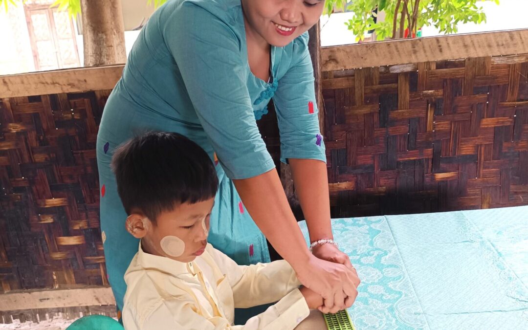7-jähriger Junge in Myanmar lernt mit Hilfe einer Lehrerin mit einer kleinen Schreibtafel Braille zu schreiben. Der Junge sitzt am Tisch und die Lehrerin ist über ihn gebeugt. Die Tischdecke ist hellblau mit weißen Blumenornamenten. Die Lehrerin hat ein hellblaues Kleid an und der Junge trägt ein beiges Hemd und einen karierten Longyi, dem traditionellen Wickelrock in Mynamar. Der Junge hat sein Thanaka-Paste im Gesicht.