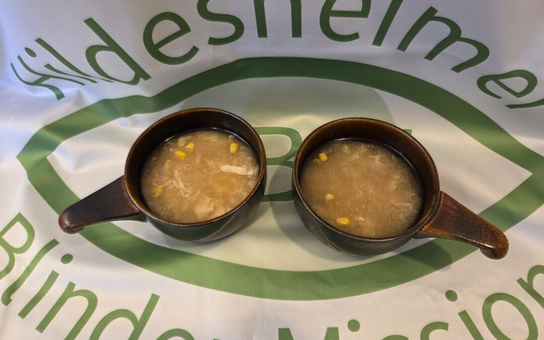 Foto: zwei kleine braune Schüsseln mit Chinesische Hühnersuppe mit Mais auf einer Tischdecke, die das Logo der Hildesheimer Blindenmission zeigt.