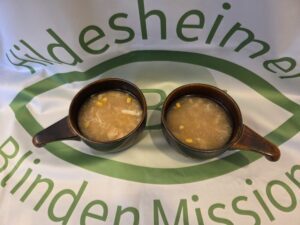 Foto: zwei kleine braune Schüsseln mit Chinesische Hühnersuppe mit Mais auf einer Tischdecke, die das Logo der Hildesheimer Blindenmission zeigt.