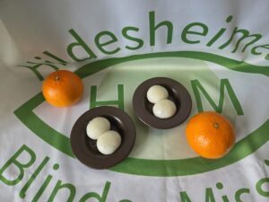 Auf einer Tischdecke mit HBM Logo stehen zwei braune Schüsseln mit je zwei weißen Klößchen drin. Außerdem liegen noch zwei Mandarinen auf der Tischdecke.