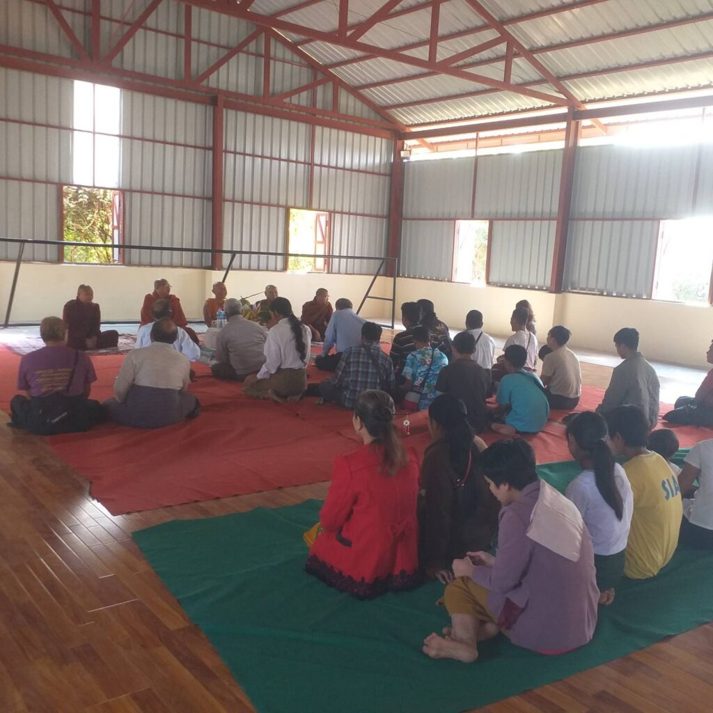 Innenansicht der Sporthalle der New Life (Bawathit) Blind School in Myanmar, Feier zur Eröffnung mit Menschen, die auf Decken sitzen, fünf Mönche sprechen ihren Segen