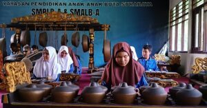 Gamelan Vorführung der Schüler und Schülerinnen in der Highschool in Surabaya. Alles sitzend an ihren Instrumenten, wie hängende und stehende Gongs, zwei große Trommeln und verschiedene Xylophone. Die Mädchen haben weiße oder rötlich braune Kopfbedeckungen. Im Hintergrund an der blauen Wand steht Yayasan pendidikan anak-anak buta Surabaya SMPLB-A & SMALB-A.