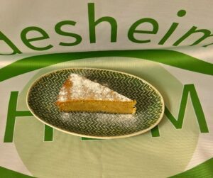 Ovaler grüner Teller mit Zickzack-Muster auf dem ein Stück Grießkuchen mit Puderzucker liegt. Der Teller steht auf einer Tischdecke mit HBM-Logo.