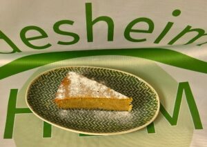 Ovaler grüner Teller mit Zickzack-Muster auf dem ein Stück Grießkuchen mit Puderzucker liegt. Der Teller steht auf einer Tischdecke mit HBM-Logo.