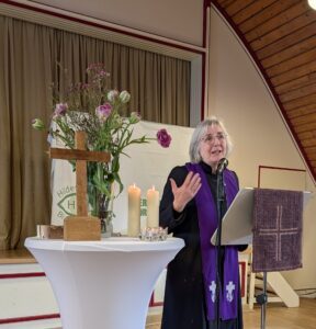 Martina Helmer‑Pham Xuan, langjährig in der Auslandsarbeit und zuletzt Pröpstin in der Ev.-luth. Landeskirche Braunschweig, hält die Predigt im Talar am Mikrofon, neben ihr ist ein Stehtisch mit Holzkreuz, Blumen und Kerzen