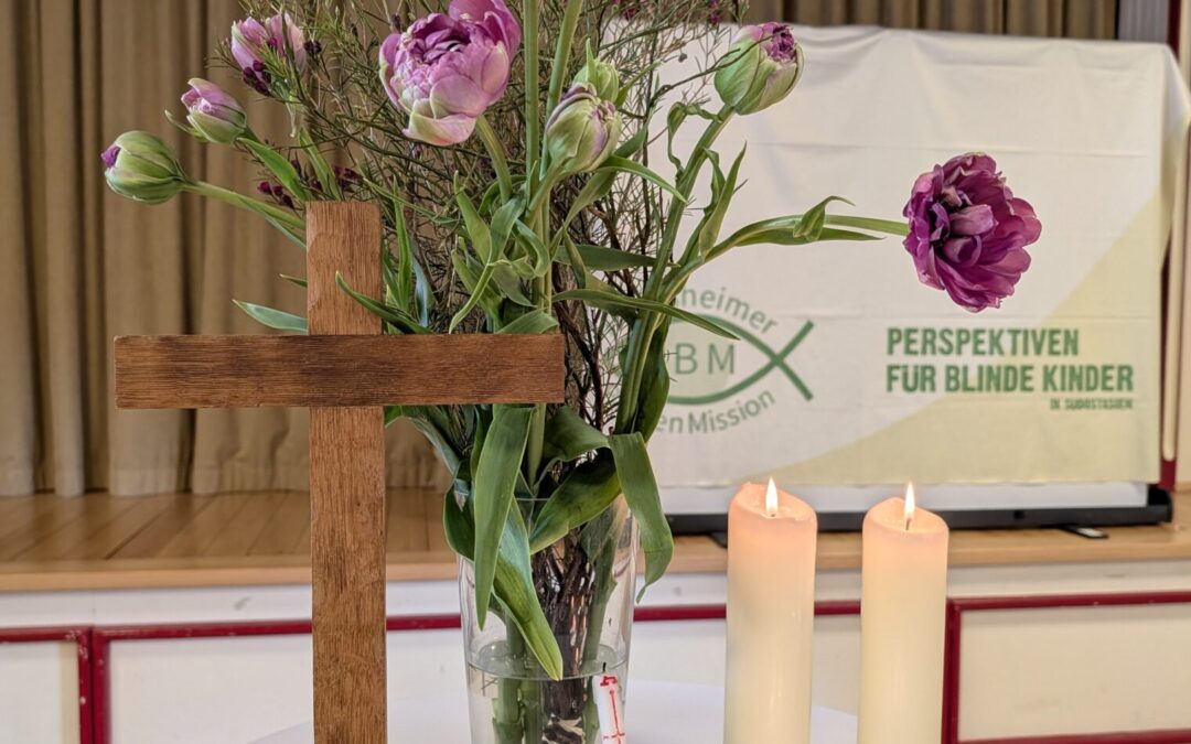 Blick auf den Stehtisch-Altar mit weißer Husse: Holzkreuz mit dickem Sockel, 2 große dicke brennende Kerzen und die drei kleinen brennenden Teelichtkerzen, die im Laufe des Godi angezündet wurden, des weiteren eine Glasvase mit einem in lila gehaltenen Blumenstrauß mit hauptsächlich gefüllten lila Tulpen, an der Vase lehnt eine kleine Kerze, die zum Anzünden der Teelichter benutzt wurde. Im Hintergrund sieht man die Bühne des Saals mit hellbraunem Vorhang und eine Leinwand, die mit einem Tuch mit HBM-Logo abgedeckt ist, auf dem Tuch steht auch: Perspektiven für blinde Kinder in Südostasien