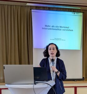Thea Hummel hält einen Vortag mit Mikrofon, vor sich hat sie auf einem Stehtisch zwei Laptops und hinter sich eine Leinwand, die das Thema des Vortrags zeigt: Mehr als ein Merkmal - Intersektionalität verstehen. Sie trägt ein weiß schwarz gestreiftes Hemd und eine blaue blazerähnliche Jacke sowie eine schwarze Armbanduhr