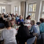 viele Menschen sitzen an Tischen und löffeln chinesische Suppe und Essen Brot dazu, die Tische sind mit einzelnen Tulpen, Butterstücke auf kleinen Tellern, Brotkörbchen, Getränken in Flaschen und Karaffen, Gläsern, Teelichtern und in der Mitte jeden Tisches ein Ulos geschmückt. Im Hintergrund ist eine Fensterreihe