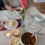 Mittagstisch mit chinesischer Suppe in Suppenschüssel mit Löffel drin auf einer Serviette mit Blätter-Ornamenten in grau weiß gehalten, daneben lieben zwei Scheiben Baguettebrot mit Butter, eine Teelichtkerze im Glashalter, ein Messer, ein halbes Stück Butter auf einer Untertasse, ein Glas Wasser, eine kleine Flasche mit einer rosa Tulpe drin, in der Tischmitte längs eine Ulos als Mitteldecke, gegenüber am Tisch sitzt eine Person ohne Kopf Suppe löffelnd, rechts an der Stirnseite steht noch eine leere Suppenschüssel auf einer Serviette mit Messer, Löffel und Glas