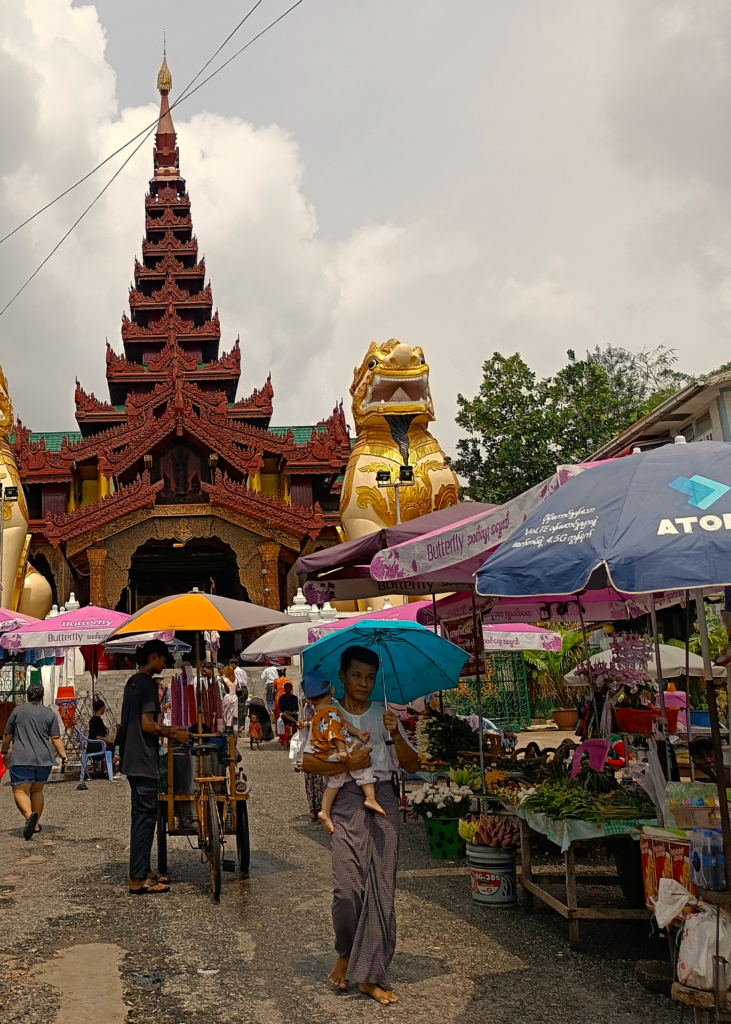 Im Fokus stehen zwei Männer vor einer großen Pagode in Myanmar. Ein Familienvater, der sein kleines Kind auf dem Arm trägt und einen blauen Schirm über sich hält. Er trägt einen Longyi und läuft barfuß. Der andere Mann hat einen dreirädrigen Fahrradstand mit gelb-grauen Schirm darüber und verkauft Eis. Er trägt T-shirt, Hose und einfache Sandalen. Weiter sind viele Sonnenschirme zu sehen und ein Blumenstand sowie Touristen kleine im Hintergrund vor der Pagode