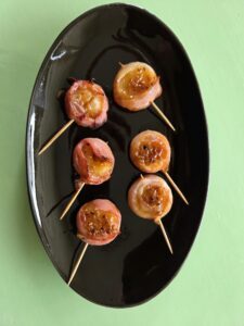 6 kleine Wasserkastanien in einem dünnen Speckmantel appetitlich glänzend von der runden Seite zu sehen mit hellem gerösteten Sesam bestreut von einem Zahnstocher zusammengehalten auf einer schwarzen glänzenden ovalen Platte wie die 6 Punkte auf einem Würfel angeordnet. Der Blick ist von oben auf die Platte mit den Wasserkastanien im Speckmantel. Die Platte steht auf einem grünen Untergrund.