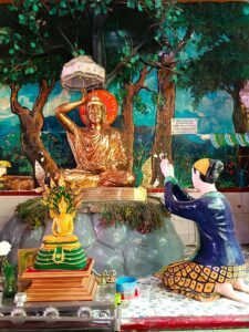 Goldener Buddha auf Steinhaufen mit weißem Schirm unter Baum im Schneidersitz sitzend. Die linke Hand mit Handfläche nach oben vor Bauch haltende, die rechte Hand über Kopf mit spezieller Fingerstellung. Der Buddha ist schlank. Vor dem Buddha kniet eine Frau mit hocherhobenen betenden Händen in Richtung des Buddhas. Sie hat hochgesteckte schwarze Haar mit gelbem kleinen Tuch, blauem Oberteil und gelb blau kariertem Rock. Sie ist barfuß. Des weiteren ist vor dem Steinhaufen ein kleinerer goldener Buddha auf einer grün goldenen Erhöhung, die auf einem rot goldenen Sockel steht, der wiederum auf einem glänzenden Metalltisch steht. Auch dieser kleine Buddha hat die typische spitze Kopfbedeckung auf. Er hält beide Hände, die locker auf dem Schoß liegen, mit dem den Handflächen nach oben. Die Erleuchtung wird auch hier mit einem Heiligenschein dargestellt. Aufgenommen wurde das Foto in Myanmar in Yangon in einer kleinen Pagode.