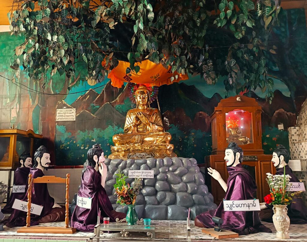 Goldener Buddha auf Steinhaufen unter Baum im Schneidersitz sitzend mit spezieller Handstellung vor der Brust, über ihm ein orangener Schirm und darüber Blätter eines Baumes, vor ihm knieend 5 diskutierende Männer in lila Umhängen. Der Buddha ist schlank. Aufgenommen wurde das Foto in Myanmar in Yangon in einer kleineren Pagode