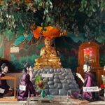 Goldener Buddha auf Steinhaufen unter Baum im Schneidersitz sitzend mit spezieller Handstellung vor der Brust, über ihm ein orangener Schirm und darüber Blätter eines Baumes, vor ihm knieend 5 diskutierende Männer in lila Umhängen. Der Buddha ist schlank. Aufgenommen wurde das Foto in Myanmar in Yangon in einer kleineren Pagode