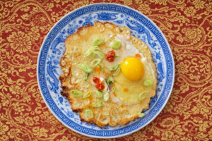 Chinesischer Pfannkuchen mit rohem Ei, Chilisauce und geschnittenen Lauchzwiebeln drauf auf einem blau mit Drachen gemusterten Teller, der auf einer roten Tischdecke mit goldenen chinesischen Blumenornamenten steht, Untergrund ist KI generiert
