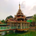 Kleine Pagode (ähnlich einem Pavillon) auf algengrünem Wasser von einem grünen Zaun umgeben. Das Dach ist rot mit vielen kleinen nach oben strebenden goldenen Verzierungen. Das Foto wurde in Myanmar in Yangon aufgenommen.