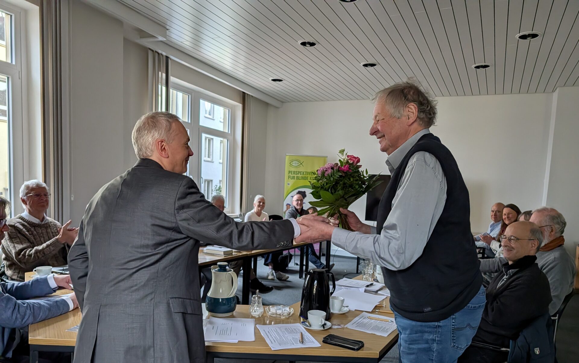 Mitgliederversammlung 2026 im großen Saal der HBM. Die Tische stehen in U-Form. Im Vordergrund übergibt Herr Chrzanowski Herrn Brinkop ein Blumenstrauß mit rosa und lila Blumen. Beide lächeln sich an. Im Hintergrund applaudieren und lächeln die restlichen anwesenden Vereinsmitglieder.