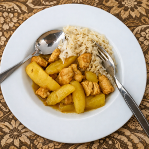 Ein weißer Teller auf einer indonesischen Tischdecke mit Löffel und Gabel. Auf dem Teller sieht man außerdem Reis, Kartoffeln, Fleisch und eine hellbraune Soße.