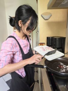 Cecilia Aloina Sembiring steht mit Schürze in der HBM-Küche am Herd und kocht Semur, ein indonesisches Schmorgericht. Gerade schupst sie geschnittene Zwiebeln von einem Brettchen mit einem Schlitzpfannenwender in eine Pfanne, die auf dem Herd steht. Das Foto ist mit KI bearbeitet.