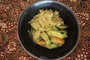 Schwarze Schüssel mit Indonesischem Duftreis und Curry-Gemüse steht auf einer indonesischen, das heißt braun, rot, gold, schwarz, weiß gemusteren Tischdecke.