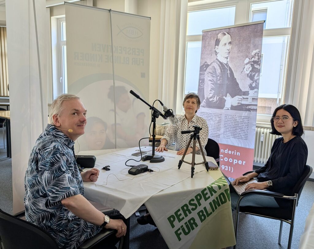 Podcastaufnahme Folge 18: Blick in die Runde mit Andreas Chrzanowski, Susanne Maier und Cecilia Sembiring. Die drei sitzen an einem Tisch mit HBM Tischdecke. Auf dem Tisch stehen Mikrofon, Stativ, Aufnahmegerät und es liegen diverse Zettel dazwischen. Umgeben ist die Dreiergruppe von HBM-Aufstellern, die das Klangerlebnis des Podcasts verbessern. Hinter Susanne Maier sieht man gut den Aufsteller mit der am Tisch-sitzenden Luise Cooper in schwarz-weiß. Andreas Chrzanowski trägt ein Headset und Cecilia Sembiring hat ihren Laptop auf dem Schoß. Alle drei schauen in die Kamera.