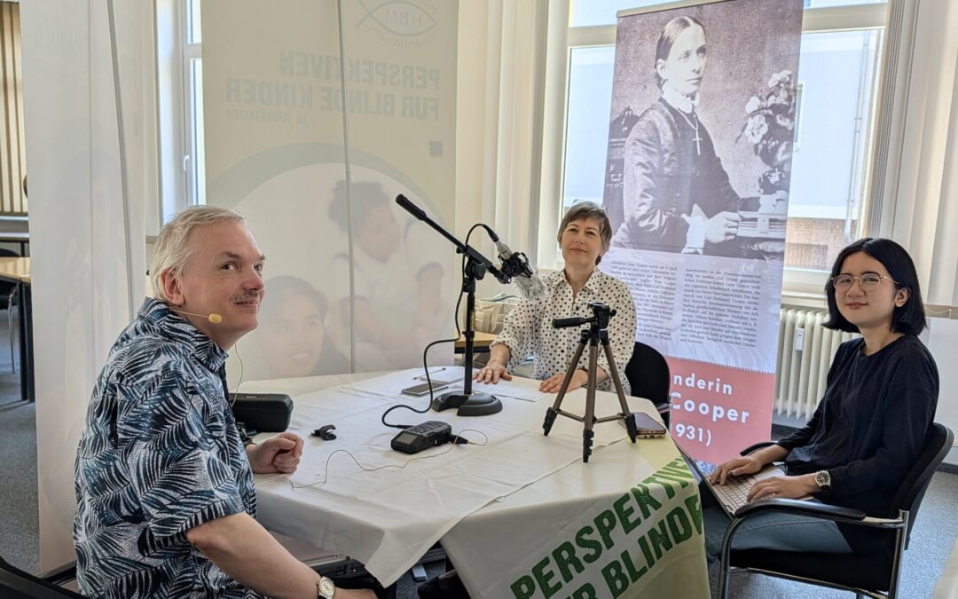 Podcastaufnahme Folge 18: Blick in die Runde mit Andreas Chrzanowski, Susanne Maier und Cecilia Sembiring. Die drei sitzen an einem Tisch mit HBM Tischdecke. Auf dem Tisch stehen Mikrofon, Stativ, Aufnahmegerät und es liegen diverse Zettel dazwischen. Umgeben ist die Dreiergruppe von HBM-Aufstellern, die das Klangerlebnis des Podcasts verbessern. Hinter Susanne Maier sieht man gut den Aufsteller mit der am Tisch-sitzenden Luise Cooper in schwarz-weiß. Andreas Chrzanowski trägt ein Headset und Cecilia Sembiring hat ihren Laptop auf dem Schoß. Alle drei schauen in die Kamera.